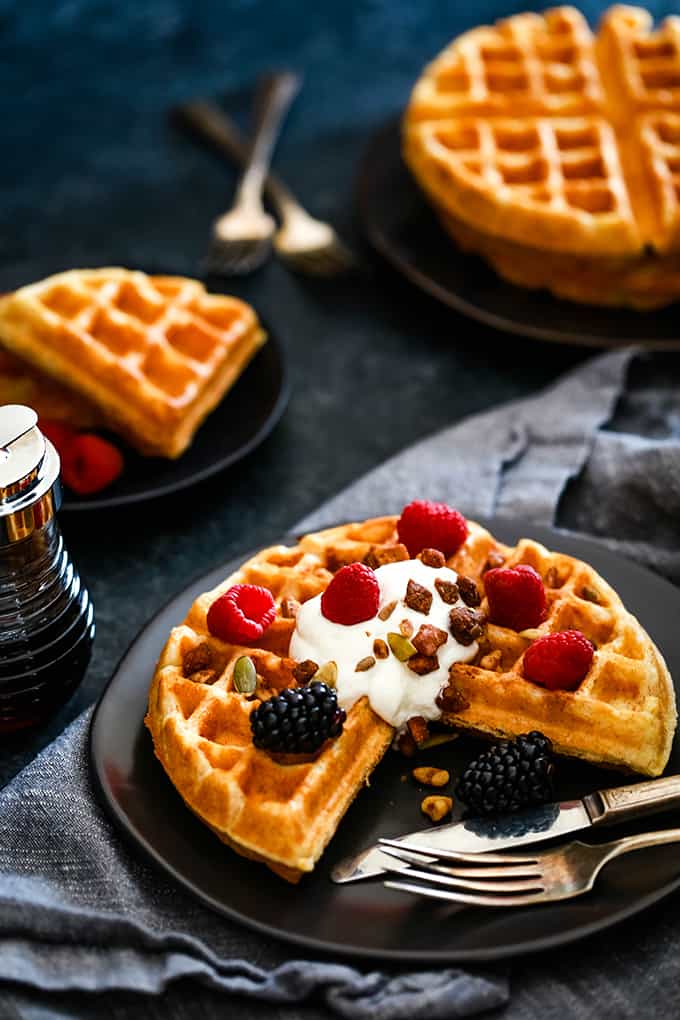 A piece of a Yogurt Waffle is missing as it sits on a dark plate with a fork and knife.