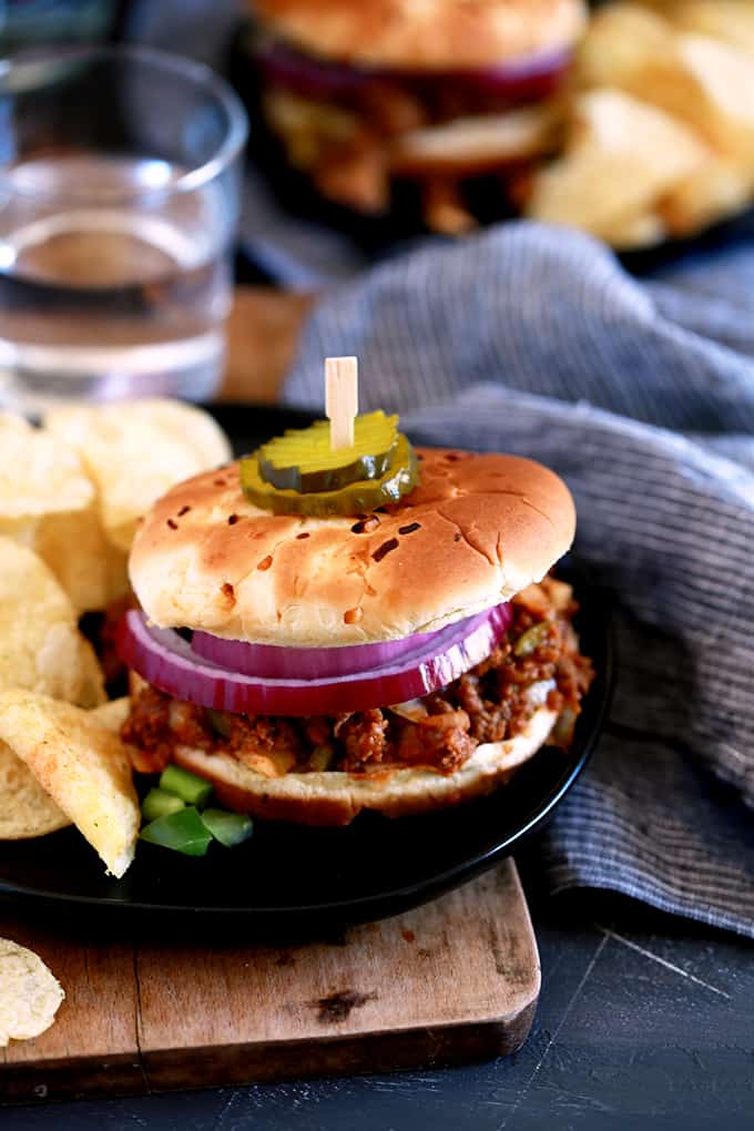 An Easy Sloppy Joes sandwich on a dark plate sits on a wooden board next to a striped napkin and water glass.