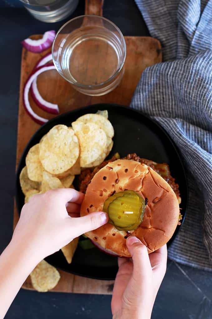 Two hands reach for an Easy Sloppy Joes sandwich off of a dark plate served with potato chips.