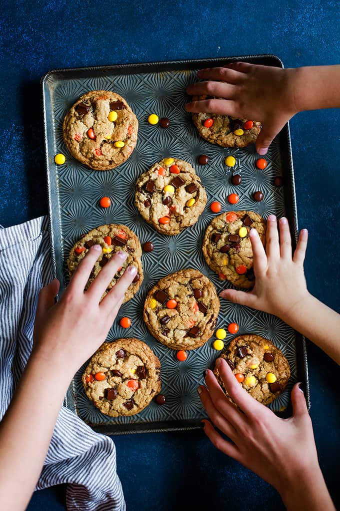 A baking sheet Reese's Pieces Chocolate Chip Cookies with four hands reaching to each take a cookie on a blue background.