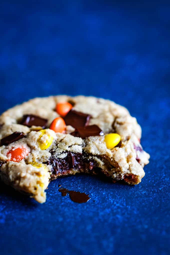 A Reese's Pieces Chocolate Chip Cookie with a bite taken out of it on a blue background.