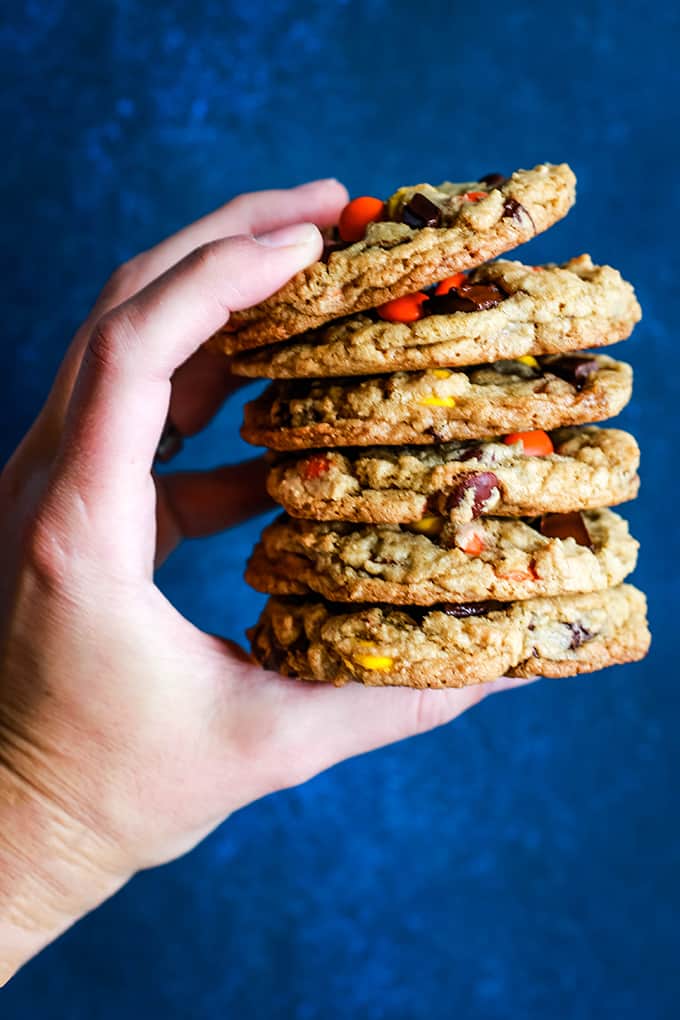 A hand holding a stack of six Reese's Pieces Chocolate Chip Cookies on a blue background.