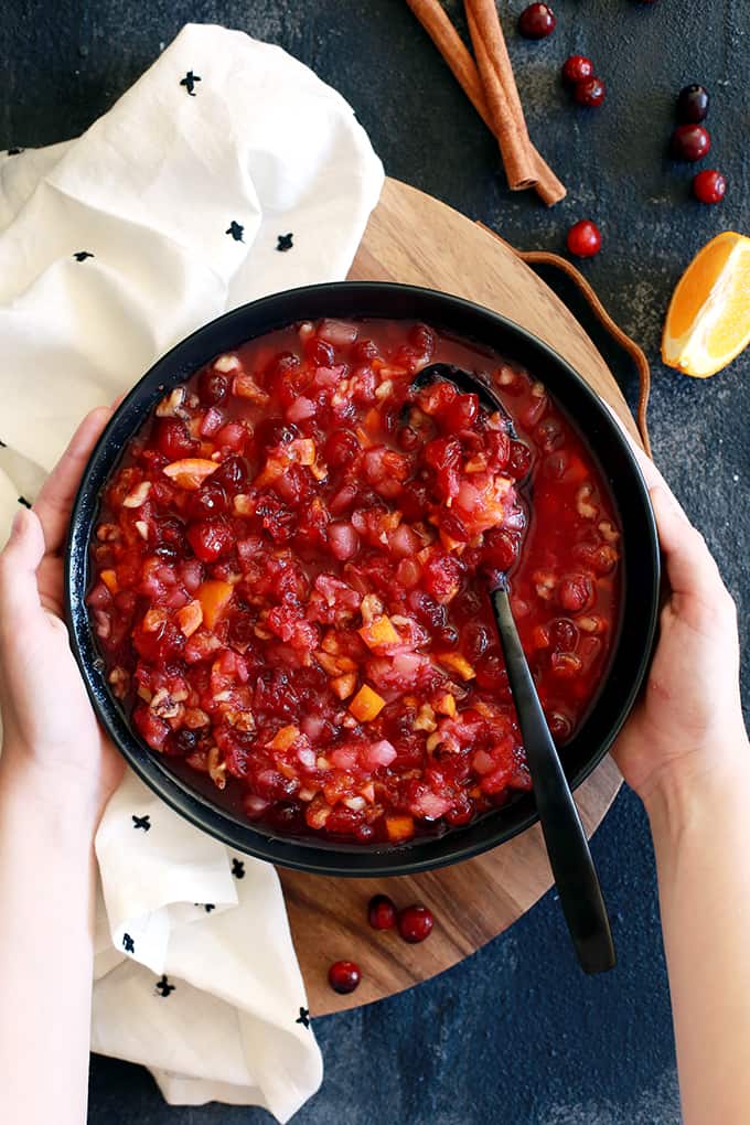 This amazing seven ingredient Citrus Cranberry Sauce is the easiest holiday side dish to grace your dinner table, with a bright and flavorful twist on an otherwise traditional dish. #cranberry #cranberrysauce #clementine #thanksgiving #thanksgivingrecipes #christmas #citrus Two hands hold a bowl of Citrus Cranberry Sauce that sits on a wooden platter and is flanked by a napkin, cinnamon sticks, cranberries and a clementine slice.
