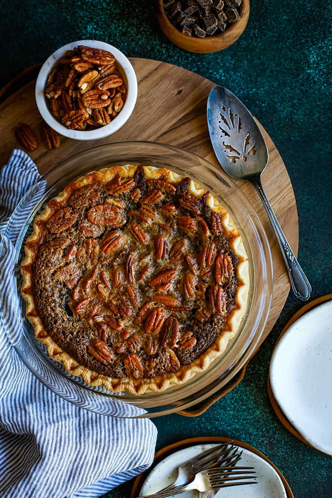 A Dark Chocolate Brownie Pie sits on a wooden tray surrounded by bowls of pecans and chocolate, a pie server, a striped napkin and plates and forks.