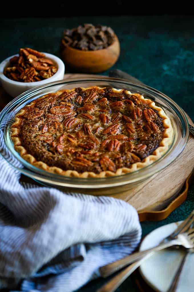 A Dark Chocolate Brownie Pie sits on a wooden tray surrounded by bowls of pecans and chocolate, a striped napkin and plates and forks.