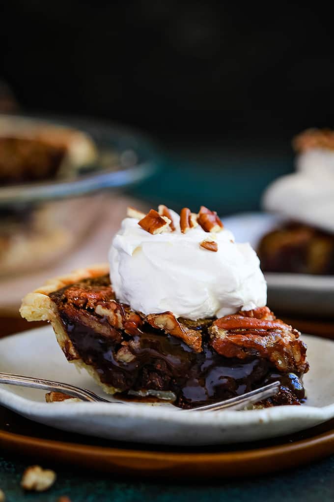 A slice of Dark Chocolate Brownie Pecan Pie sits on a white plate with a fork on a wooden charger.