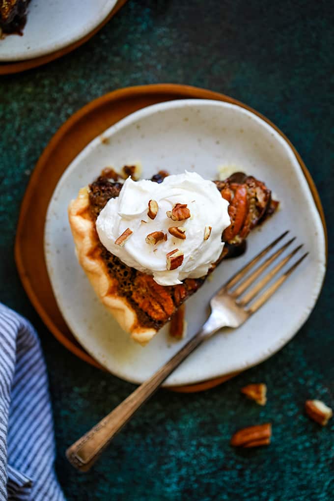 An overhead shot of a slice of Dark Chocolate Brownie Pecan Pie sitting on a white plate on a wooden charger with a fork on the plate and surrounded by a striped napkin, additional pecans and a second plate.