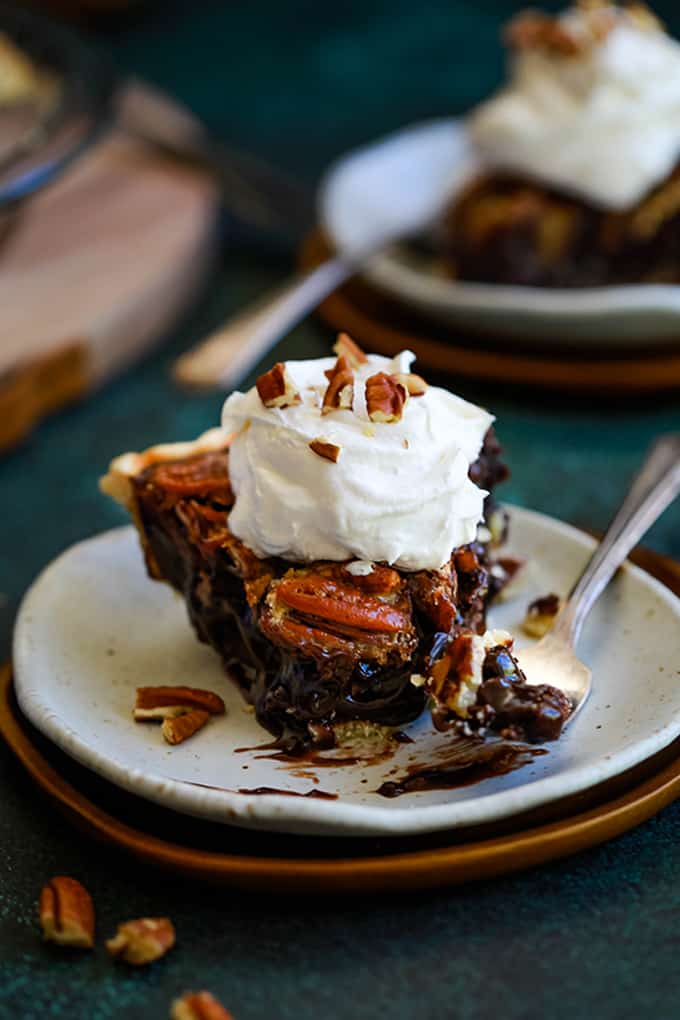 A slice of Dark Chocolate Pecan Pie sits on a white plate with a bite removed. The plate since in front of another plate and a wooden tray.