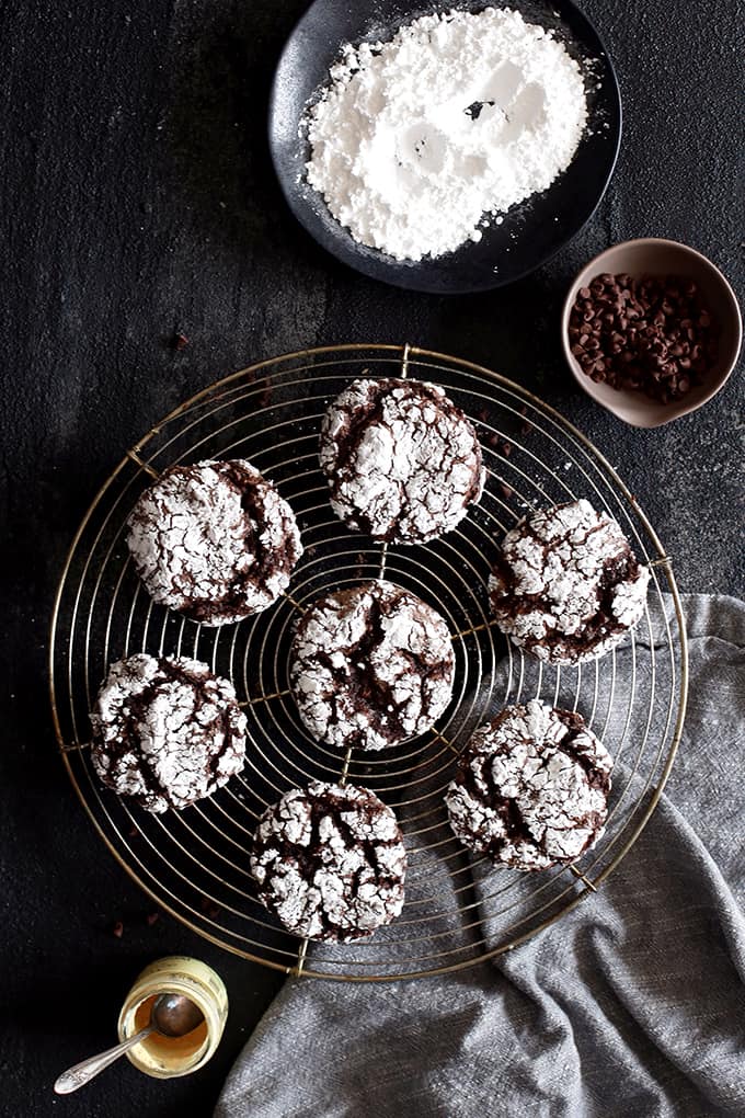 Double Chocolate Ginger Crinkle Cookies rest on a wire cooling rack surrounded by bowls of powdered sugar, chocolate chips and ginger and a grey napkin.