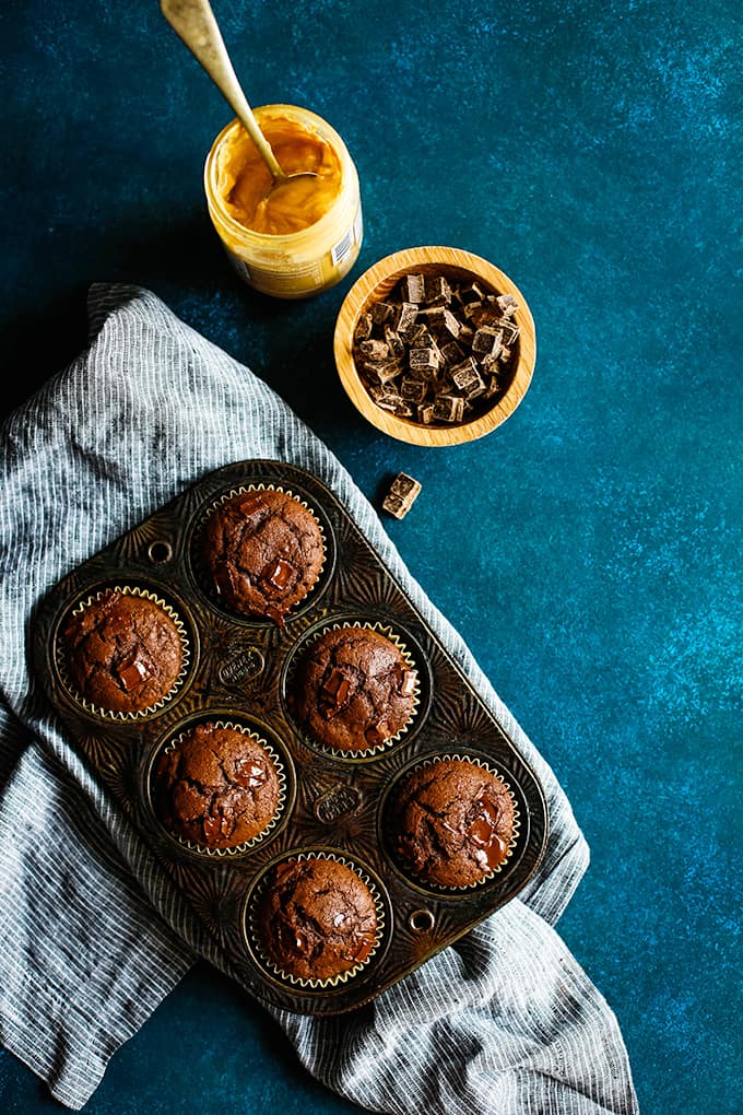 A muffin tin of Peanut Butter Mocha Chocolate Chip Muffins rests on a striped napkin surrounded by a bowl of chocolate chunks and a jar of peanut butter.