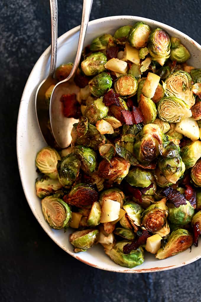 An overhead shot of a bowl of Roasted Brussels Sprouts with Apple and Bacon with two spoons on a dark background.