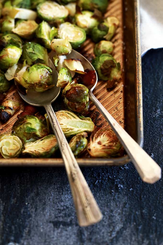 A sheet pan of Brussels sprouts for Roasted Brussels Sprouts with Apple and Bacon with two spoons sits on a dark background.