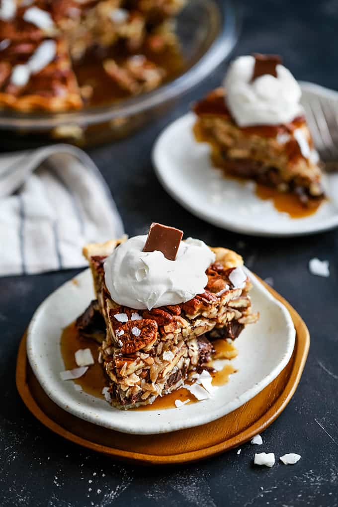 A slice of German Chocolate Pecan Pie sits on a white plate on a wooden charger that is surrounded by a striped napkin, an additional pie slice on a white plate and a pie plate.