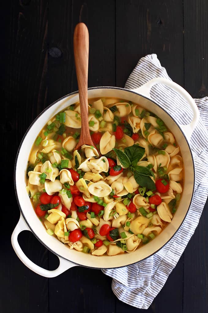 A white pot of Cherry Tomato and Tortellini Soup with a wooden spoon, flanked by a striped napkin.
