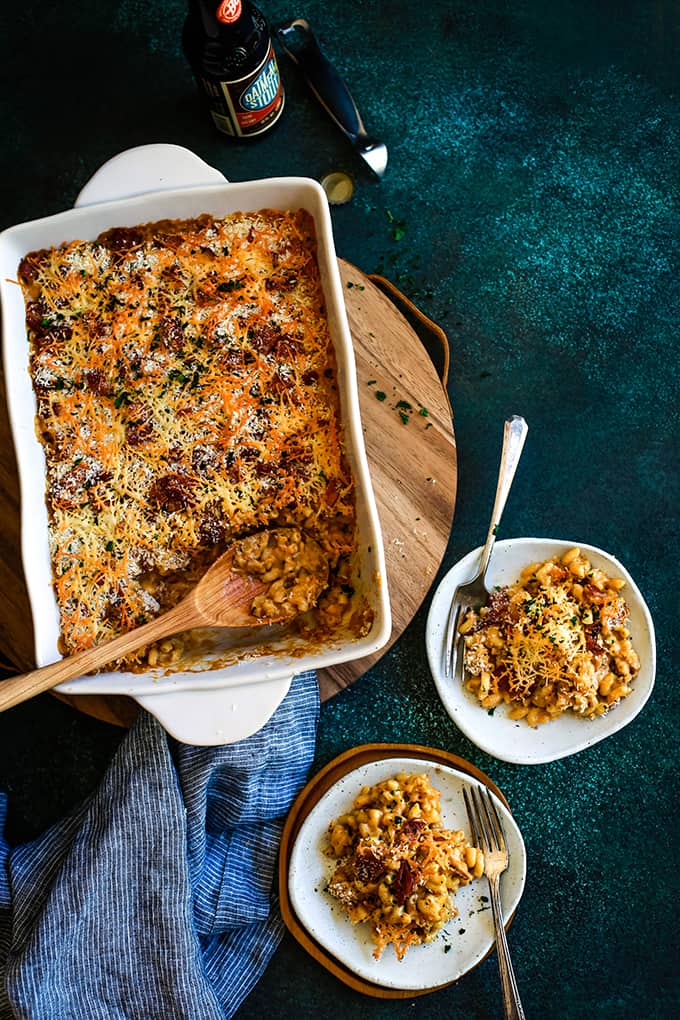 A baking dish of Bacon Crusted Beer Mac and Cheese sits on a wooden tray alongside two white plates.
