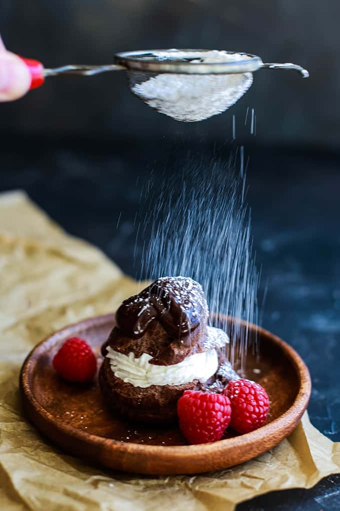 Chocolate Cream Puff on a wooden plate, filled with whipped cream and topped with chocolate ganache and powdered sugar. Garnished with fresh raspberries. A hand is sifting powdered sugar which is showing over the cream puff.