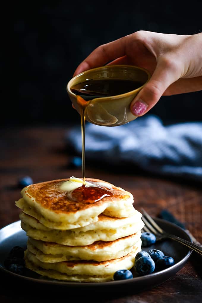 A hand pours syrup onto a stack of the Best Buttermilk Pancakes.