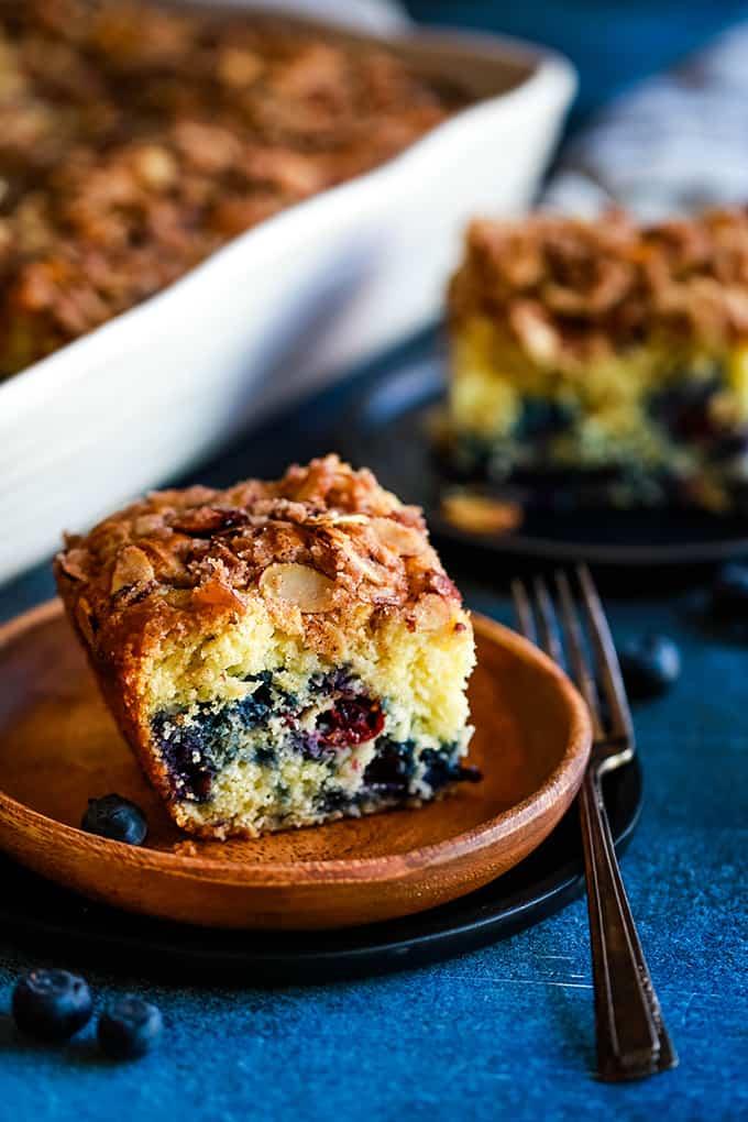 A slice of Blueberry Lemon Almond Coffee Cake on a wooden plate in front of a casserole dish.