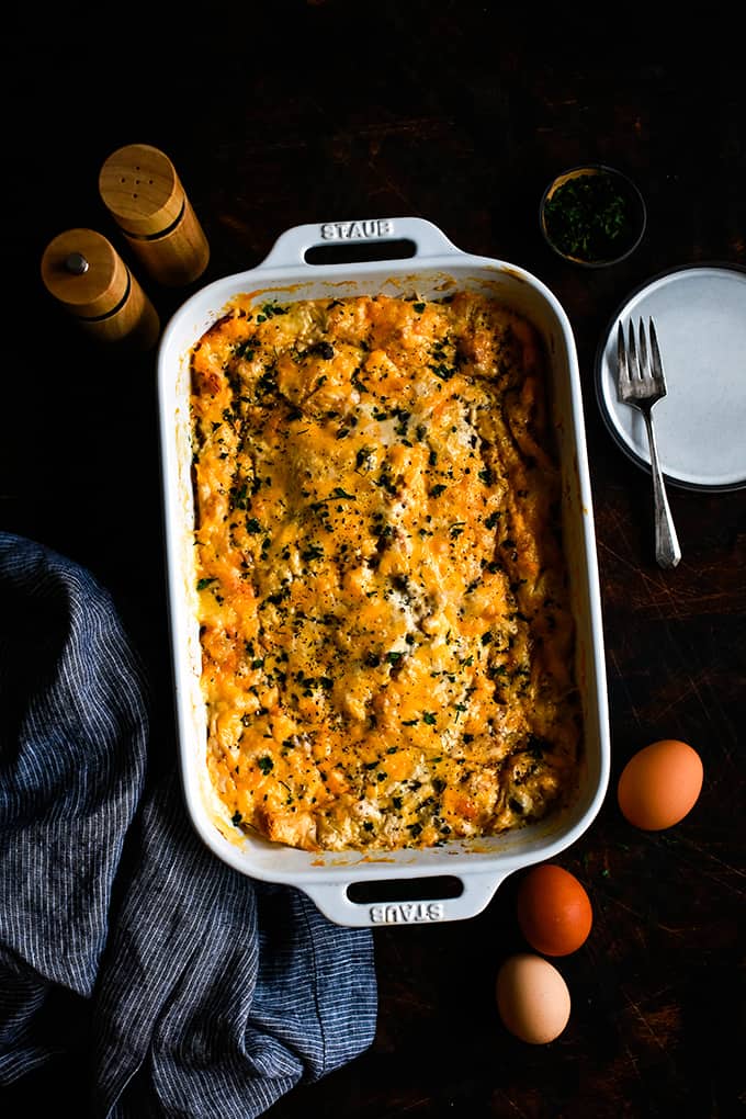 A baking dish of Bridge Brunch Casserole surrounded by a striped napkin, plate, eggs and salt and pepper shaker.