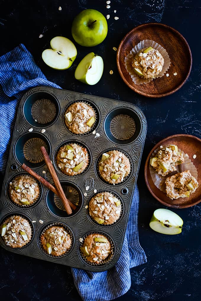 Cinnamon Apple Oatmeal Muffins in a muffin tin with a striped towel on a black background. Two wooden plates each hold a muffin and apples are alongside.