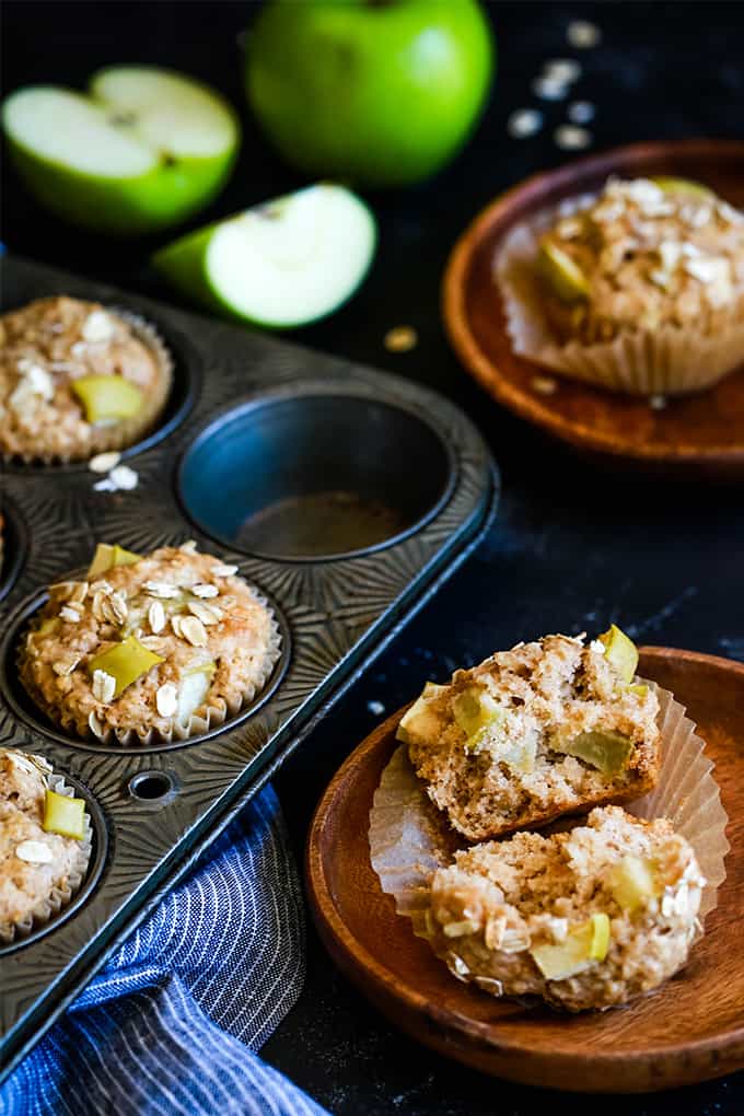 Cinnamon Apple Oatmeal Muffins in a muffin tin with a striped towel on a black background. Two wooden plates each hold a muffin and apples are alongside.