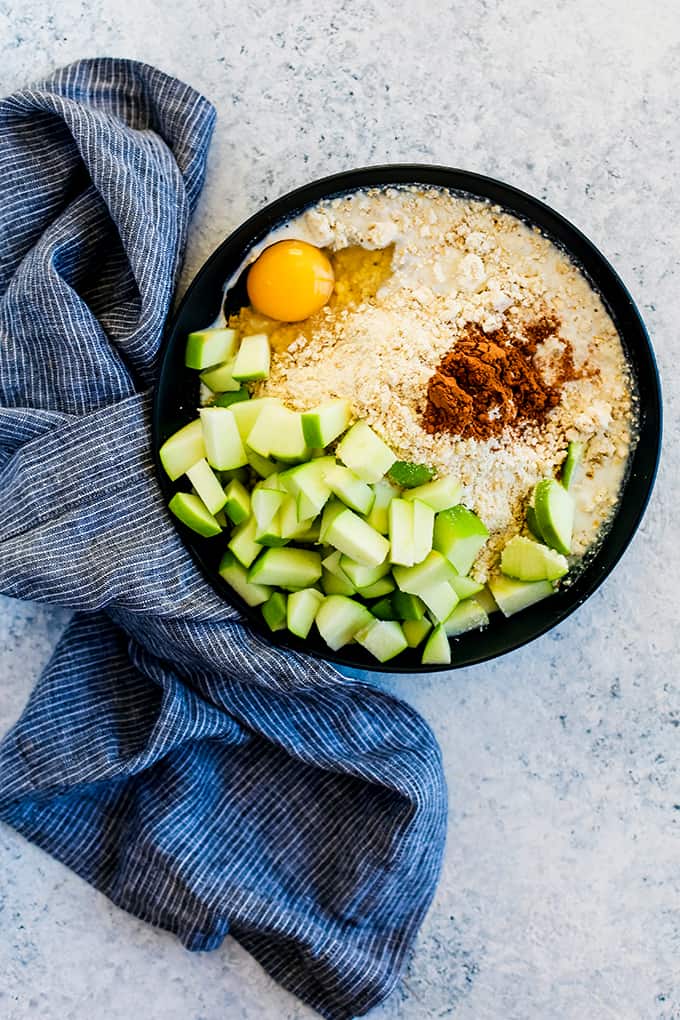 Homemade Oatmeal Muffin Mix, diced apples, ground cinnamon, egg and milk in a black bowl with a striped towel on a white and grey background.