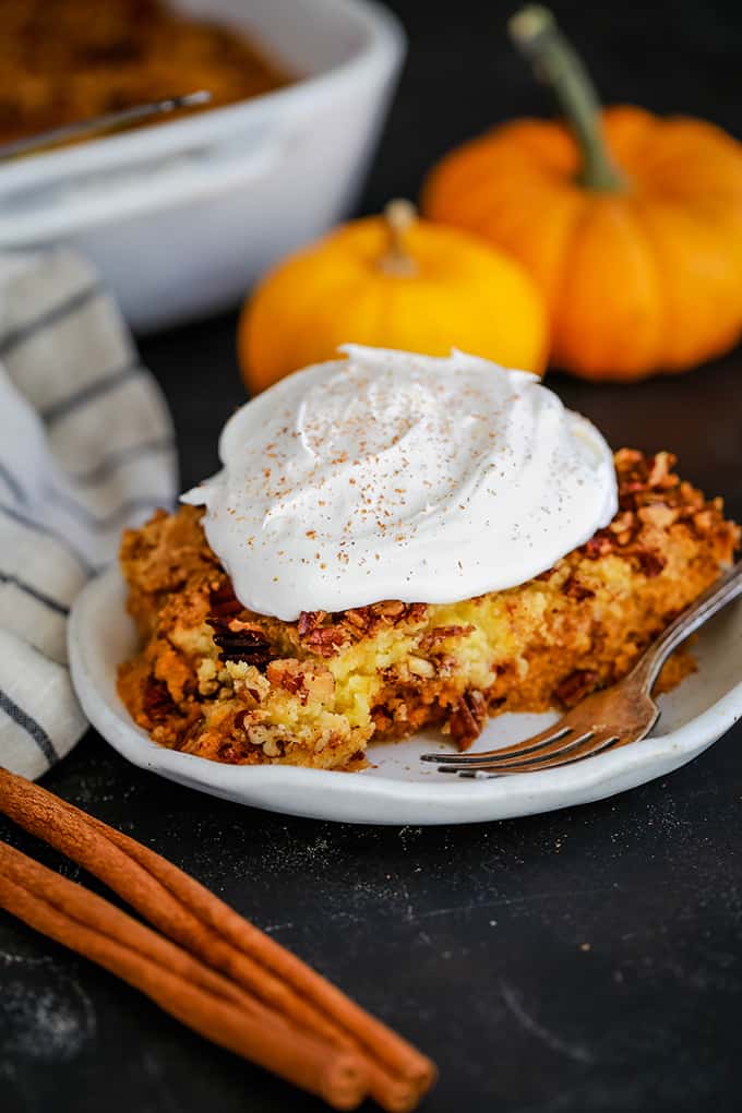 A white plate of Upside Down Pumpkin Pie topped with whipped topping is surrounded by cinnamon sticks, a striped napkin, two mini pumpkins and a white casserole dish.