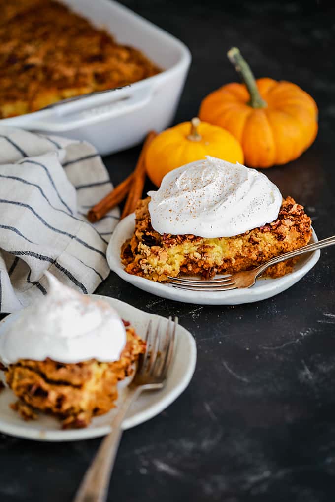 Two white plates of Upside Down Pumpkin Pie topped with whipped topping are surrounded by two mini pumpkins, a striped napkin and a serving dish.