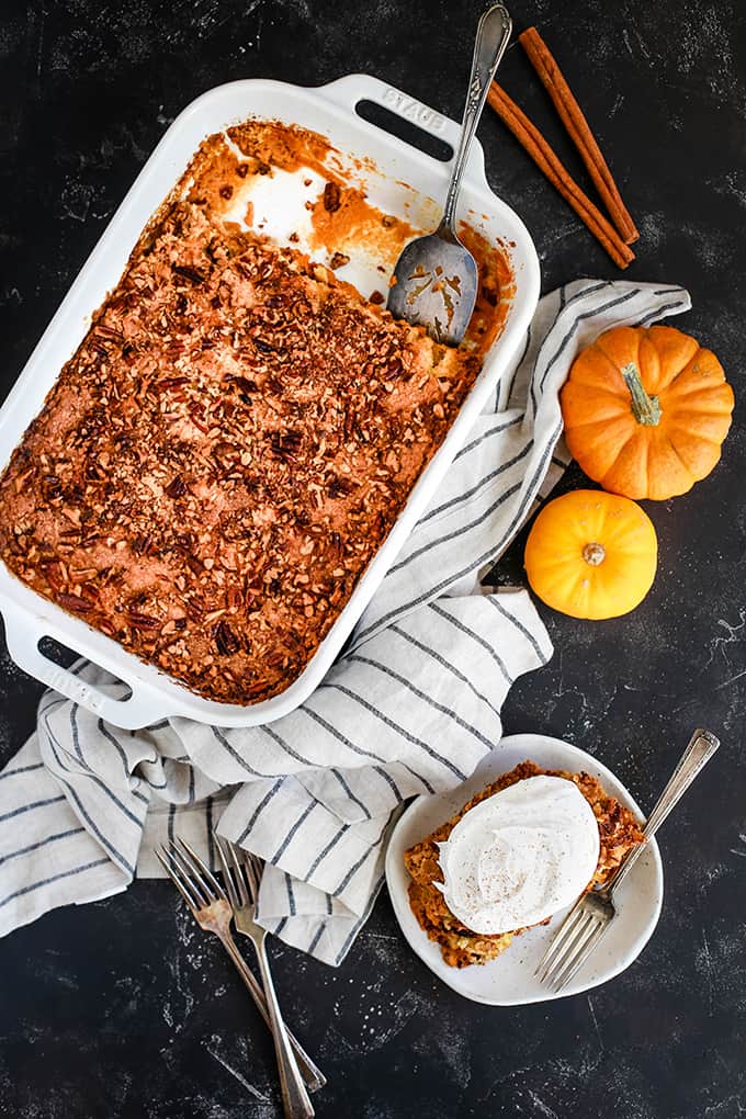 A pan of Upside Down Pumpkin Pie sits on a dark background surrounded by a striped napkin, cinnamon sticks, two mini pumpkins and a white plate with Upside Down Pumpkin Pie topped with whipped topping.