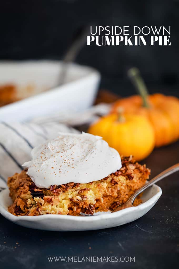 A slice of Upside Down Pumpkin Pie sits on a white plate with a fork on a dark surface. A striped napkin and two mini pumpkins are in the background.