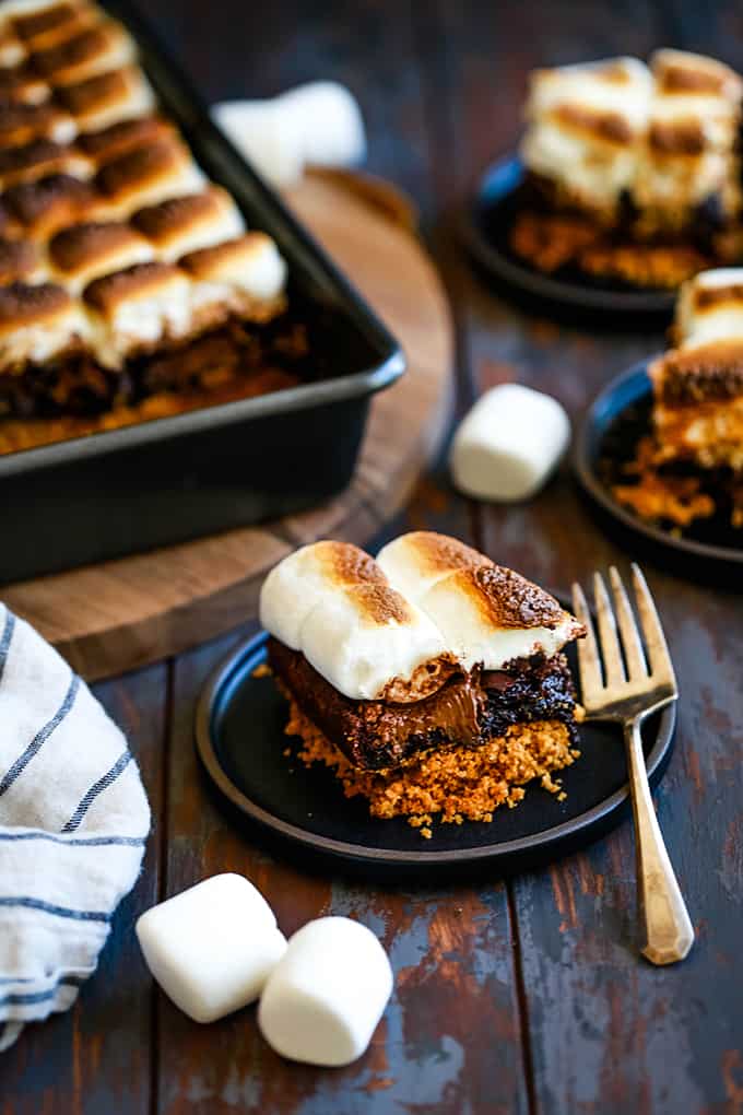A Peanut Butter Cup Stuffed S'mores Brownie sits on a black plate with other brownies in the background.