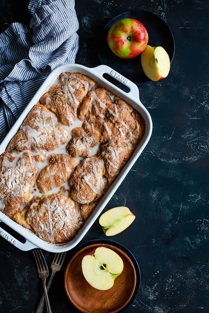 An overhead shot of a baking dish of Caramel Apple CheesecakeÂ Coffee Cake sits on a black background surrounded by a wooden plate and a black plate with cut apples, a striped napkin and two forks.