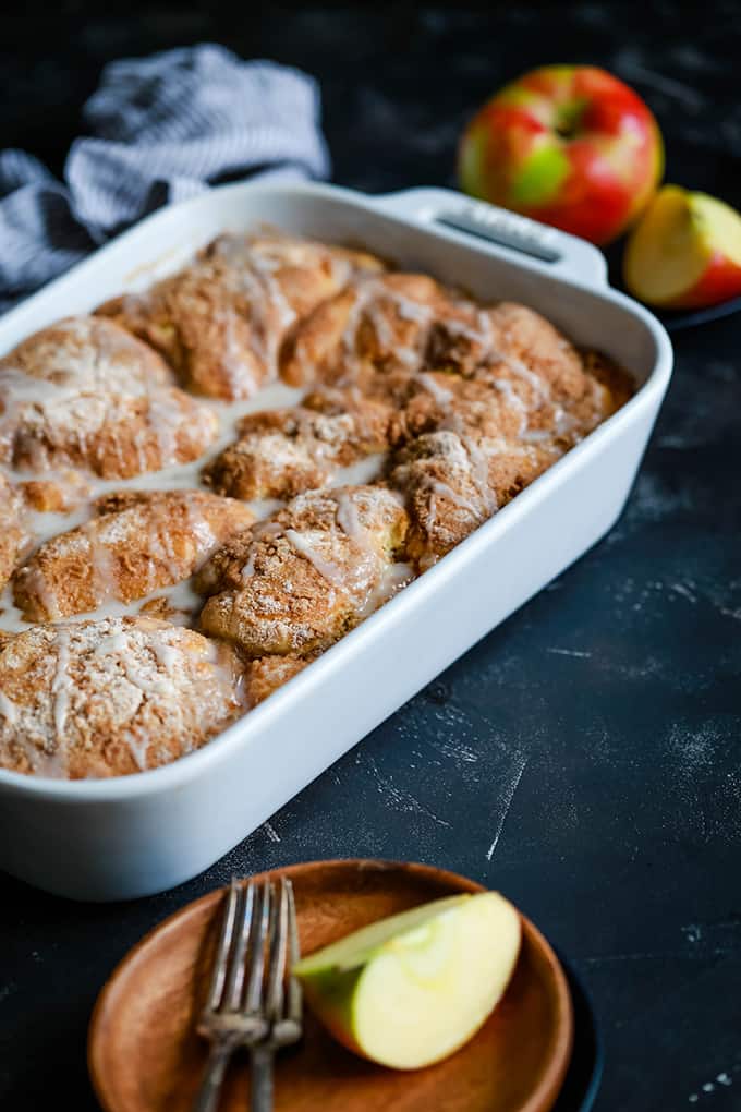 A baking dish of Caramel Apple Cheesecake Coffee Cake sits on a black background surrounded by a wooden plate and a black plate with cut apples, a striped napkin and two forks.