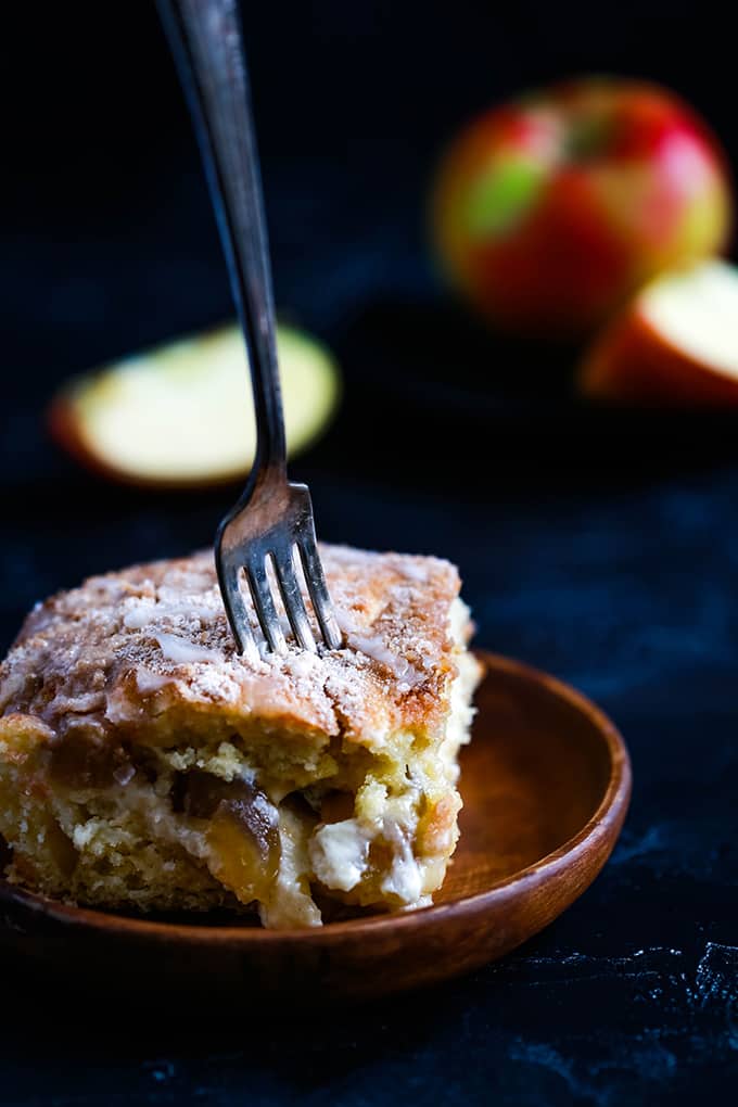 A slice of Caramel Apple CheesecakeÂ Coffee Cake on a wooden plate with a fork piercing the top. Cut apples sit in the background.