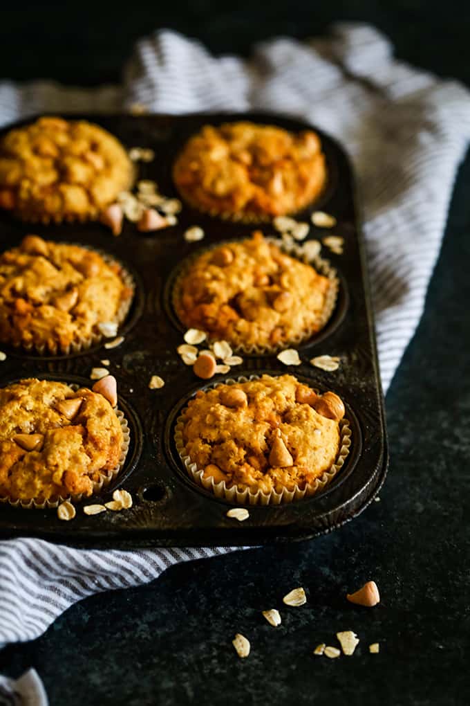 A muffin tin of Pumpkin Butterscotch Oatmeal Muffins sits on a striped napkin on a dark background.