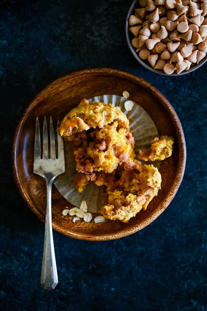 A wooden plate holds a split Pumpkin Butterscotch Oatmeal Muffin on a dark background. A smaller bowl of butterscotch chips sits in the upper right hand corner.