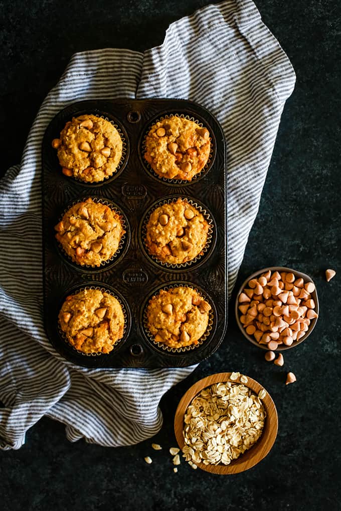 A muffin tin of Pumpkin Butterscotch Oatmeal Muffins sits on a striped napkin on a dark background with a small wooden bowl of dry oatmeal and a smaller bowl of butterscotch chips.