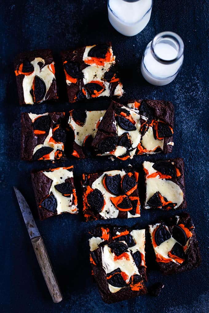 Squares of Oreo Cheesecake Brownies sits on a black background along with a knife and two bottles of milk.