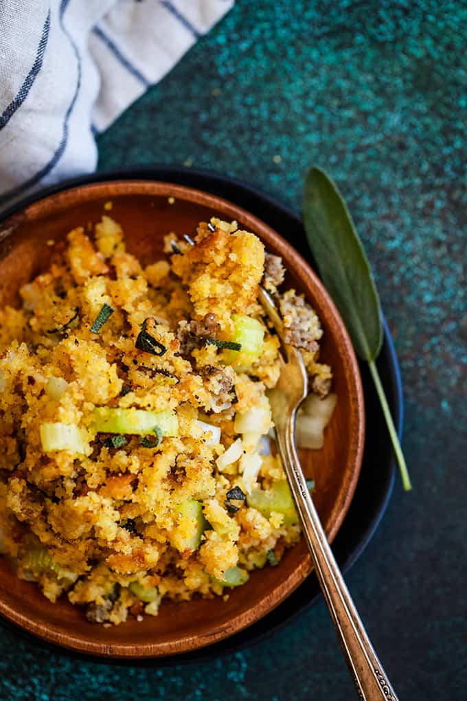 A wooden bowl full of Slow Cooker Cornbread and Sausage Stuffing sits on a black and green speckled background with a fork and a sage leaf on the side, flanked by a striped napkin.