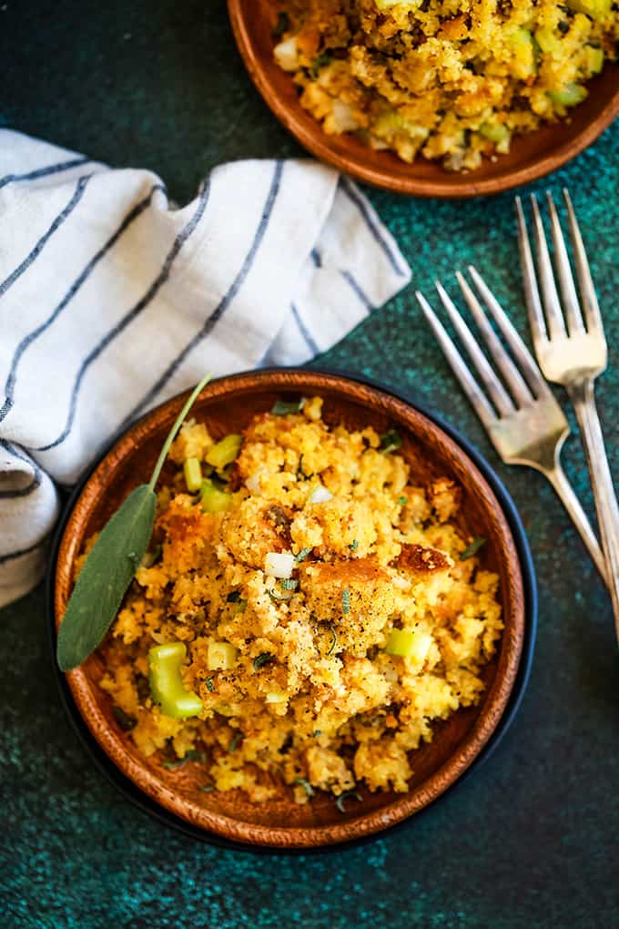 Two wooden plates full of Slow Cooker Cornbread and Sausage Stuffing sit on a black and green speckled background flanked by a striped napkin and two forks.