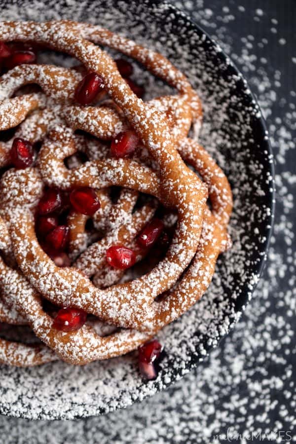 These Mini Eggnog Funnel Cakes are sure to be the star of any holiday party or gathering. A cinnamon eggnog batter is fried into a web of interlacing circles before being topped by a snow shower of powdered sugar and studded with pomegranate arils (seeds) for the perfect pop of holiday color. 