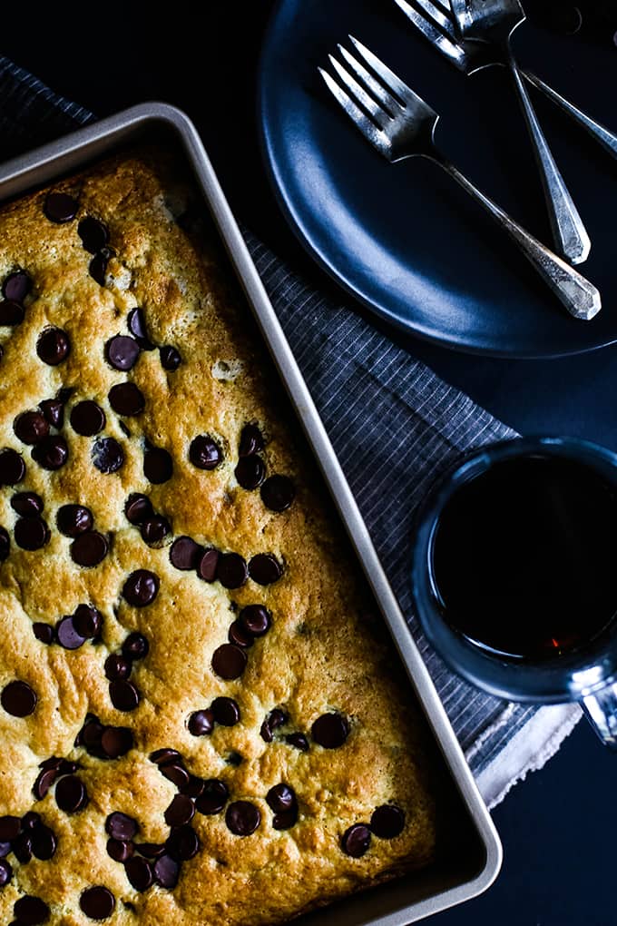 A pan of Chocolate Chip Sour Cream Coffee Cake sitting on a striped napkin with a cup of coffee, black plate and forks.