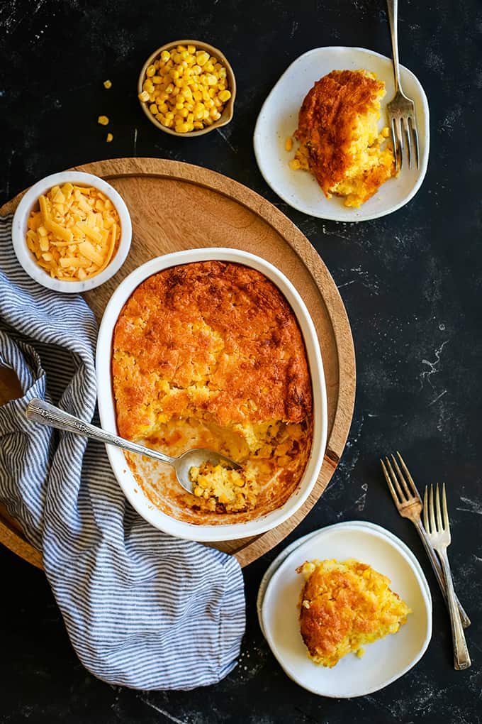 An overhead shot of a dish of Cheesy Corn Casserole with a spoon sitting on a wooden tray surrounded by two white plates full of casserole, forks, a striped napkin and two small bowls holding corn and cheese.