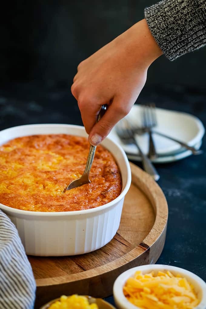 A hand inserts a spoon into a white dish of Cheesy Corn Casserole on a wooden platter, surrounded by white plates and forks and two small bowls full of cheese and corn.