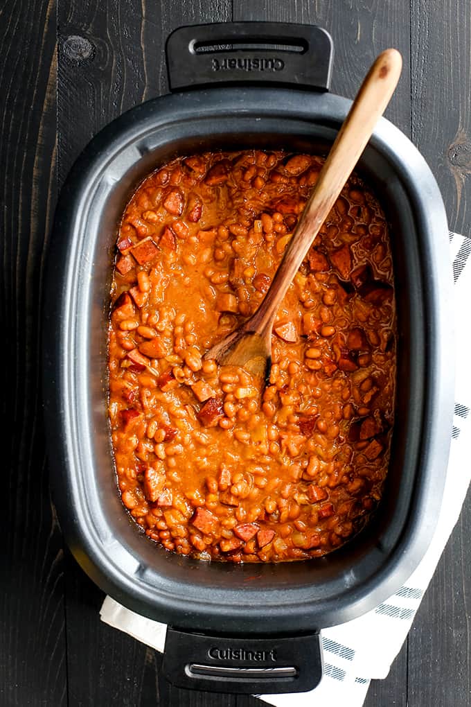 A slow cooker insert of Slow Cooker Smoky Baked Beans and a wooden spoon.