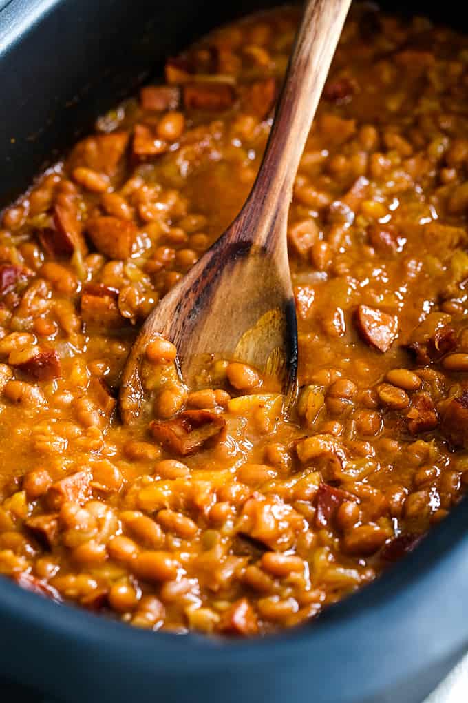 A wooden spoon rests in a slow cooker filled with Slow Cooker Smoky Baked Beans.