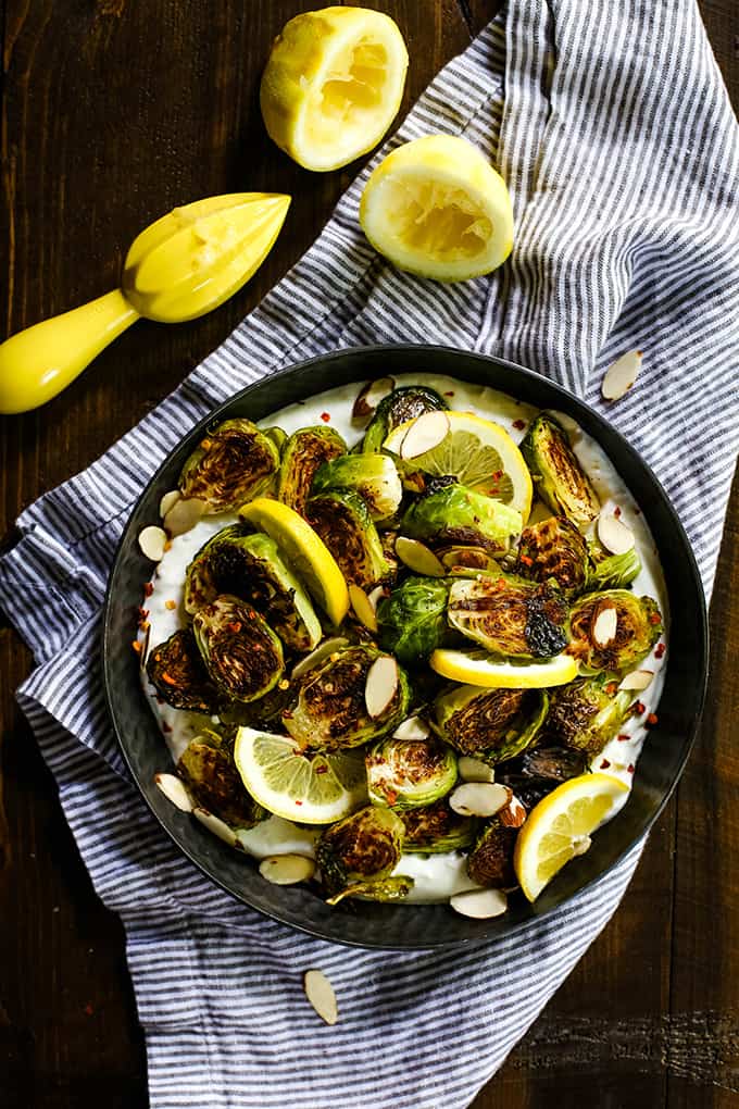 A bowl of Roasted Brussels Sprouts with Lemon and Greek Yogurt on a striped napkin flanked by two halves of a lemon.