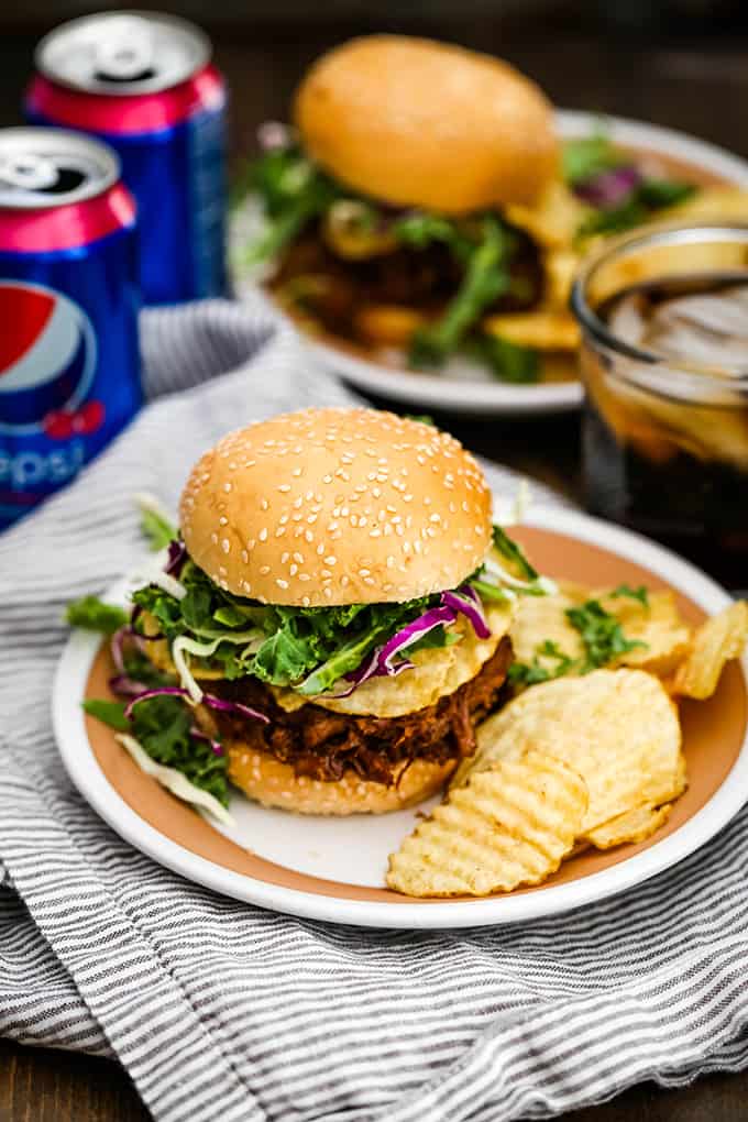 Two Slow Cooker Cherry Cola Pulled Pork Sandwiches are surrounded by cans of soda.