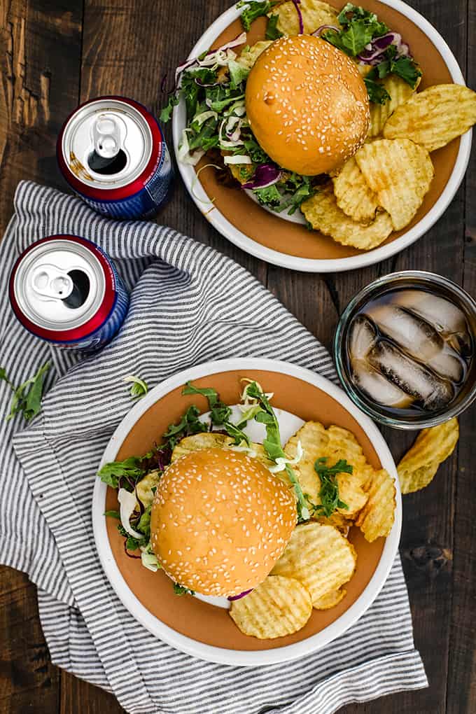An overhead view of two plates of Slow Cooker Cherry Cola Pulled Pork Sandwiches with two cans of soda and a striped napkin.