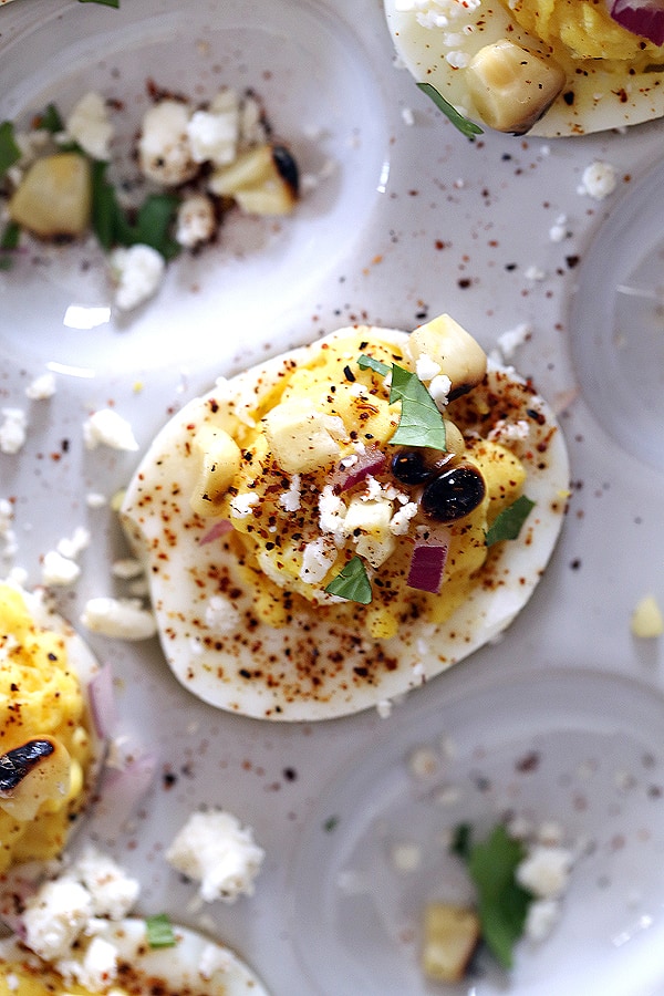 These Mexican Street Corn Deviled Eggs are guaranteed to be the talk of your next gathering! Traditional deviled eggs are garnished with feta cheese, grilled corn, red onion, chili powder and cilantro to create one amazing bite size appetizer.